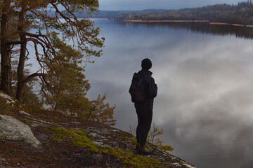Fototapeta premium A man in dark hiking gear standing on a rocky cliff, looking out over a calm, misty lake at dawn. A peaceful, atmospheric scene of solitude and Scandinavian nature during a moody autumn morning.