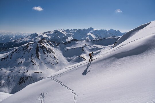 Skier Traversing Vast Snowy Alpine Slope, Lone Backcountry Guide Skiing Across Windswept Plateau Toward Jagged Peaks, Crisp Blue Sky, Ski tour to the Hochfinsler mountain with a view of the Pizol.