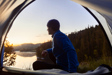 A young man in a blue fleece and beanie sits at a tent opening, gazing at a serene lake and forest during a golden sunrise. Perfect shot for outdoor adventure, camping, and peaceful nature travel. © Francis