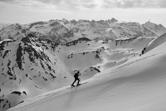 Skier Traversing Vast Snowy Alpine Slope, Lone Backcountry Guide Skiing Across Windswept Plateau Toward Jagged Peaks, Crisp Blue Sky, Sharp Shadows, Visible Tracks, Highaltitude.