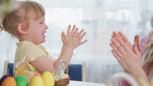 Happy child claps hands with smiling mother during Easter celebration. Mother and daughter playfully interact with colorful eggs on table. Scene captures joyful