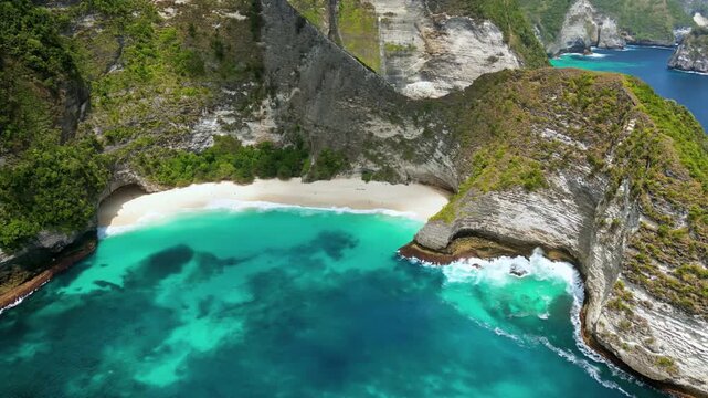 Aerial drone view of Kelingking Beach in Nusa Penida, Bali, Indonesia. Dramatic limestone cliffs tower above a pristine white sand beach and vibrant turquoise water. The famous T-Rex shaped headland d
