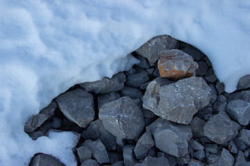 Snow-covered rocky terrain with scattered gray stones and a prominent orange rock partially exposed beneath a layer of white snow © Yahor