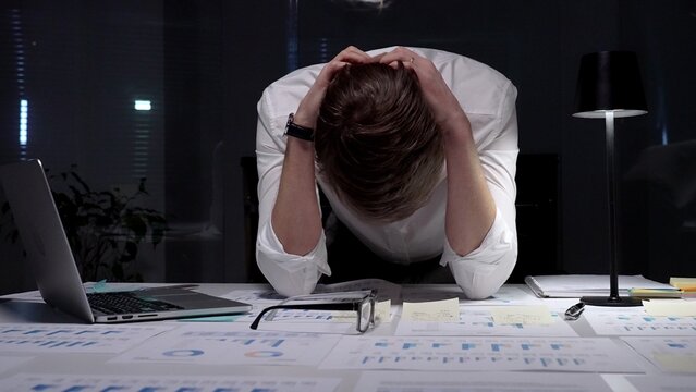 Stressed businessman suffering from overwhelming burnout and work overload, working late into the night, feeling frustrated with deadline pressure in a dark office environment