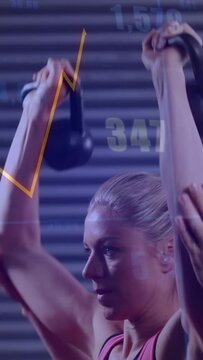 Vertical video: Trainee pressing KBs overhead from rack, coach spotting, to track fitness metrics