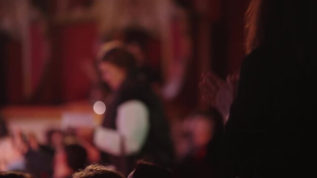 A person in a dark, atmospheric venue claps their hands in applause. The focus is on the motion of clapping, with the blurred audience and warm lighting creating an immersive feel