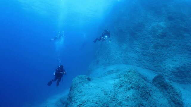  scuba divers exploring around a reef underwater deep blue water big rocks and bubbles ocean scenery 