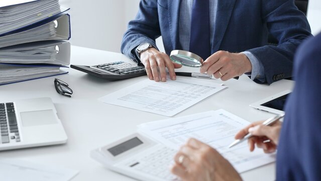 Male financial analysts with female colleague examining documents, using magnifying glass and calculator at workplace in office. Audit and taxes in business