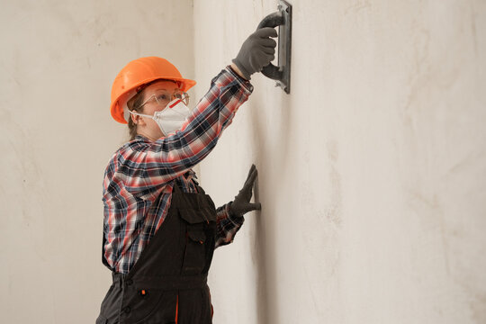 Builder woman sanding and smoothing walls after plastering for leveling getting it ready for painting during home renovation and construction work.