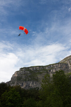 Skydiver with orange canopy soaring over the green forest and cliffs of Sarca Valley.
