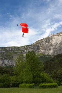 BASE jumper with vibrant orange parachute landing in a green meadow beneath the Italian Alps.