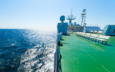 Naklejka premium Empty passenger deck of large ferry ship sailing on deep blue sea under clear sky. White benches stand on green floor of ferry near navigation equipment and radar domes