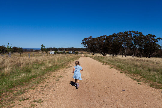Little farm girl under blue summer sky running down gravel road through paddocks