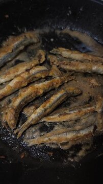 Small breaded fish, likely smelt or capelin, frying in hot, bubbling oil in a black cast iron pan for a delicious seafood meal