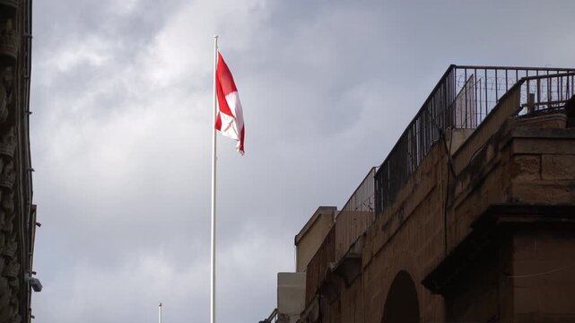 A flag with red and white triangles waves in the wind in old town Valletta, Malta. Cloudy sky is in behind.