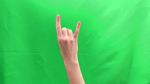 Female hand showing horns gesture with two fingers against green chroma background. Woman forming rock music sign used in concerts and metal culture. Entertainment symbol concept for fan expression.