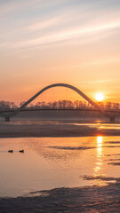 Obraz premium Golden sunrise over the modern pedestrian bridge across the Dnipro River in Kyiv, Ukraine. Calm water, winter riverbanks and silhouettes create a peaceful morning city landscape.
