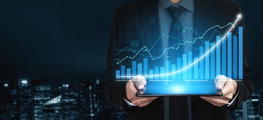 A businessman stands in a futuristic office at night, holding a tablet displaying vibrant graphs and data visualizations that signify financial growth and analysis. Impute © InfiniteFlow