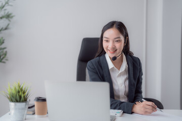 Fototapeta premium Smiling Asian businesswoman in a headset and suit working on a laptop at her office desk, providing virtual customer support during a video conference and online meeting