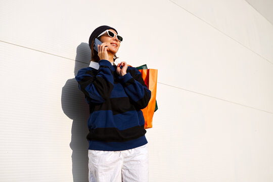 Young korean woman wearing sunglasses talking on smartphone while holding colorful shopping bags outdoors against minimal wall