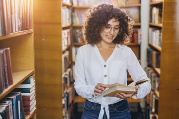 Woman with curly hair and glasses standing in a library, smiling and reading a book between bookshelves © anatoliycherkas
