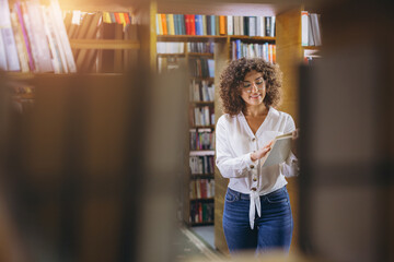 Woman standing in a library, holding an open book, and reading. She is smiling, surrounded by bookshelves filled with books © anatoliycherkas