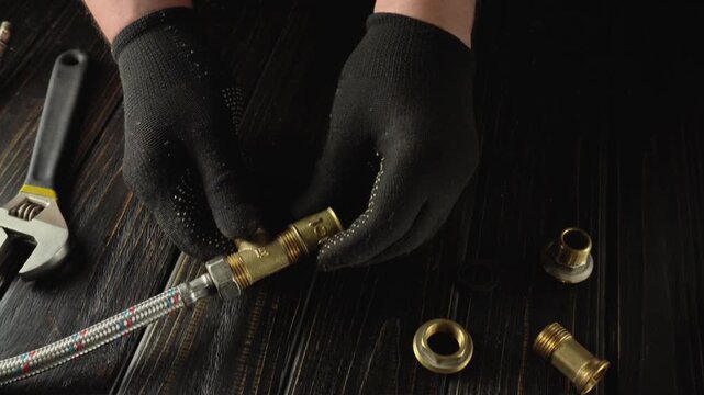 Hands in black gloves connect brass fittings and a braided hose while assembling plumbing components on a dark wooden work surface