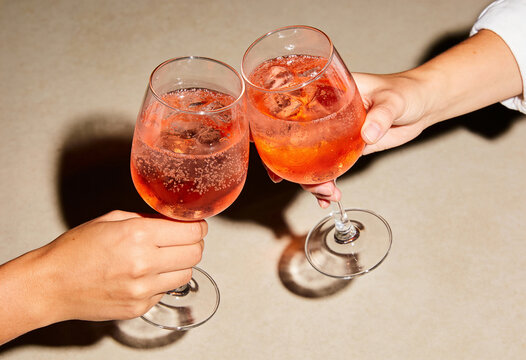 Two hands clinking glasses of orange spritz cocktail in a toast. Refreshing aperitivo with ice and bubbles on a neutral table. Social celebration and summer drinks concept