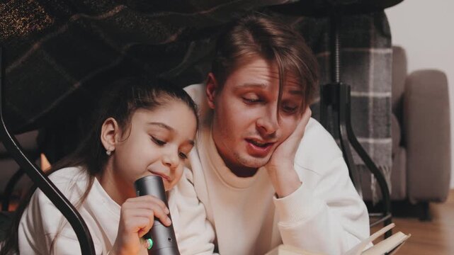 Father and daughter read story under blanket fort with flashlight in cozy indoor setting during evening
