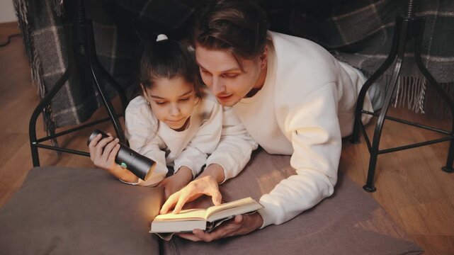 Father and daughter read together with flashlight in a cozy blanket fort at home during evening time