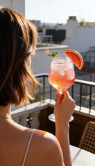 Fototapeta premium Woman holding a pink spritz cocktail on a sunny balcony. Vertical photo of a refreshing summer drink with grapefruit and mint overlooking city rooftops. Lifestyle and travel concept