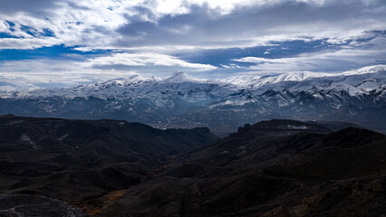 Fototapeta premium Aerial view of majestic snow-capped mountain peaks and dark rugged valleys under a dramatic cloudy sky
