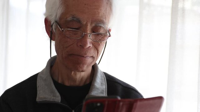 Slow motion of senior Japanese man with glasses using smartphone at home, cinematic lifestyle, silver generation, farsightedness, focused expression, elderly, 4K 30p, natural light.
