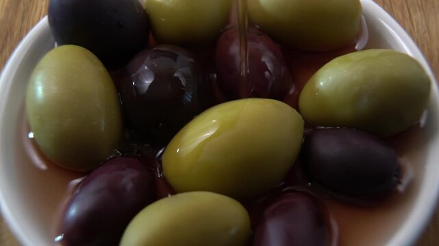 Pouring brine over assorted Olives (Olea europaea), liquid dynamics on the waxy cuticle, industrial wet curing and canning visual.