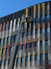 A worker on an elevated boom lift platform performs maintenance on the colorful corrugated metal facade of a curved contemporary building on a construction site