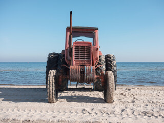 weathered, vintage red tractor stands parked on a sandy beach directly facing the calm blue ocean