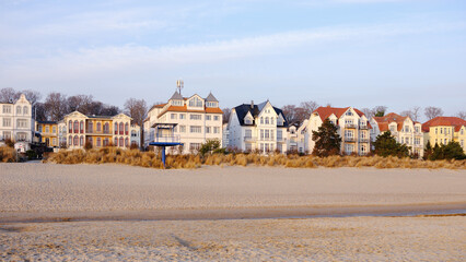 A row of classic white resort architecture and a modern blue lifeguard tower sit behind sand dunes in Bansin, Usedom, Germany