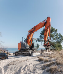 A large orange crawler excavator sits on a sandy beach dune with pine trees and clear blue sky in the background, suggesting coastal construction or erosion control work