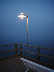 Fototapeta premium A glowing street lamp stands at the end of a wooden pier in dense blue fog, with a white bench in the foreground and calm water merging into the misty horizon