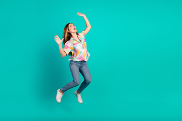 Young woman with long hair joyfully jumping in casual colorful shirt and jeans against a vibrant teal background