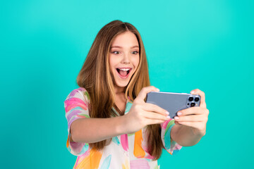 Happy young woman taking a selfie on a smartphone against teal background, showcasing summer style and joyful expression