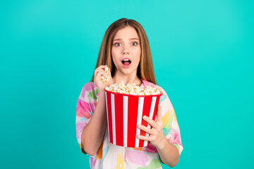 Young woman enjoying popcorn from a striped bucket, expressing surprise on a bright turquoise background