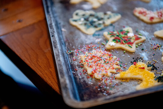 An aged cookie sheet holds raw Christmas cutout cookies covered in messy sanding sugar, the uneven application revealing a child's enthusiastic hand in the decorating.