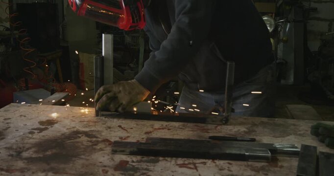 Skilled worker in protective gear using a welding machine to join metal components, creating bright sparks and smoke in an industrial garage environment while crafting a custom steel frame.