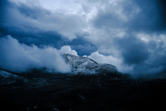 Cinematic moody shot of a dark snow-capped mountain peak emerging from heavy dramatic rolling clouds and thick fog