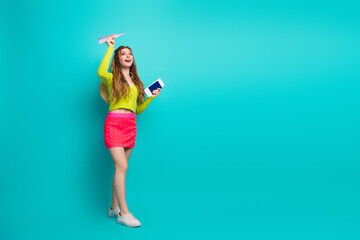 Energetic young woman in vibrant outfit holding a paper plane and booklet, expressing happiness against a turquoise background.