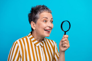 Senior woman with short gray hair smiles while examining with a magnifying glass against a bright...
