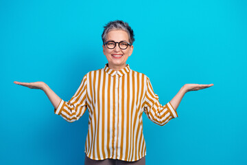 Aged female senior model in striped shirt stands with arms open smiling against blue background © deagreez