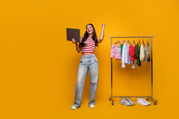 Young woman with laptop posing beside clothing rack smiling in bright studio yellow background