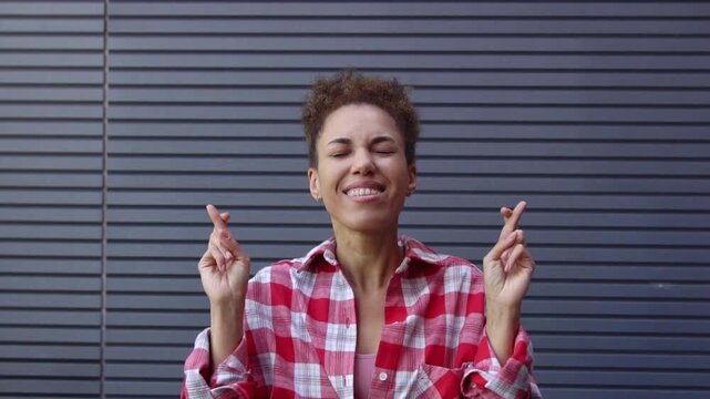 African American woman hopes and believes, crossing her fingers against urban wall, girl asks for good luck and fortune outdoors